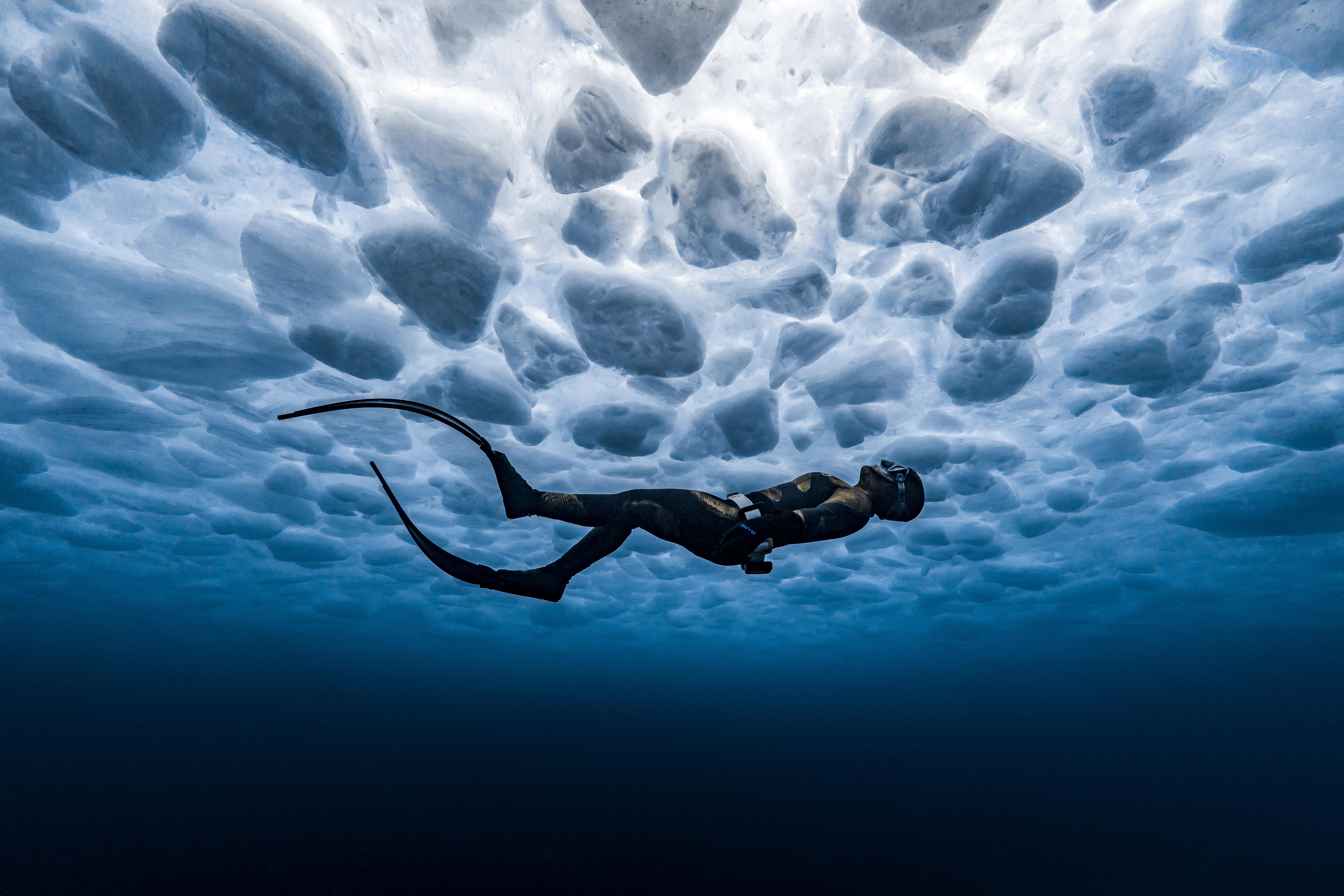 A freediver gazes up at the intricate ice patterns below the surface of a frozen lake. Canada